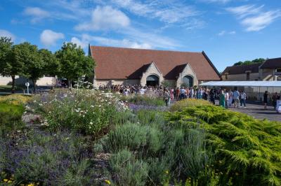 Un bâtiment en pierre avec un toit en tuiles rouges, entouré de jardins fleuris, accueille un groupe de personnes.