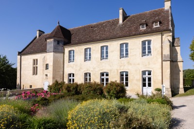 Façade d'un bâtiment ancien en pierre avec toit en ardoise, entouré d'un jardin fleuri sous un ciel bleu.