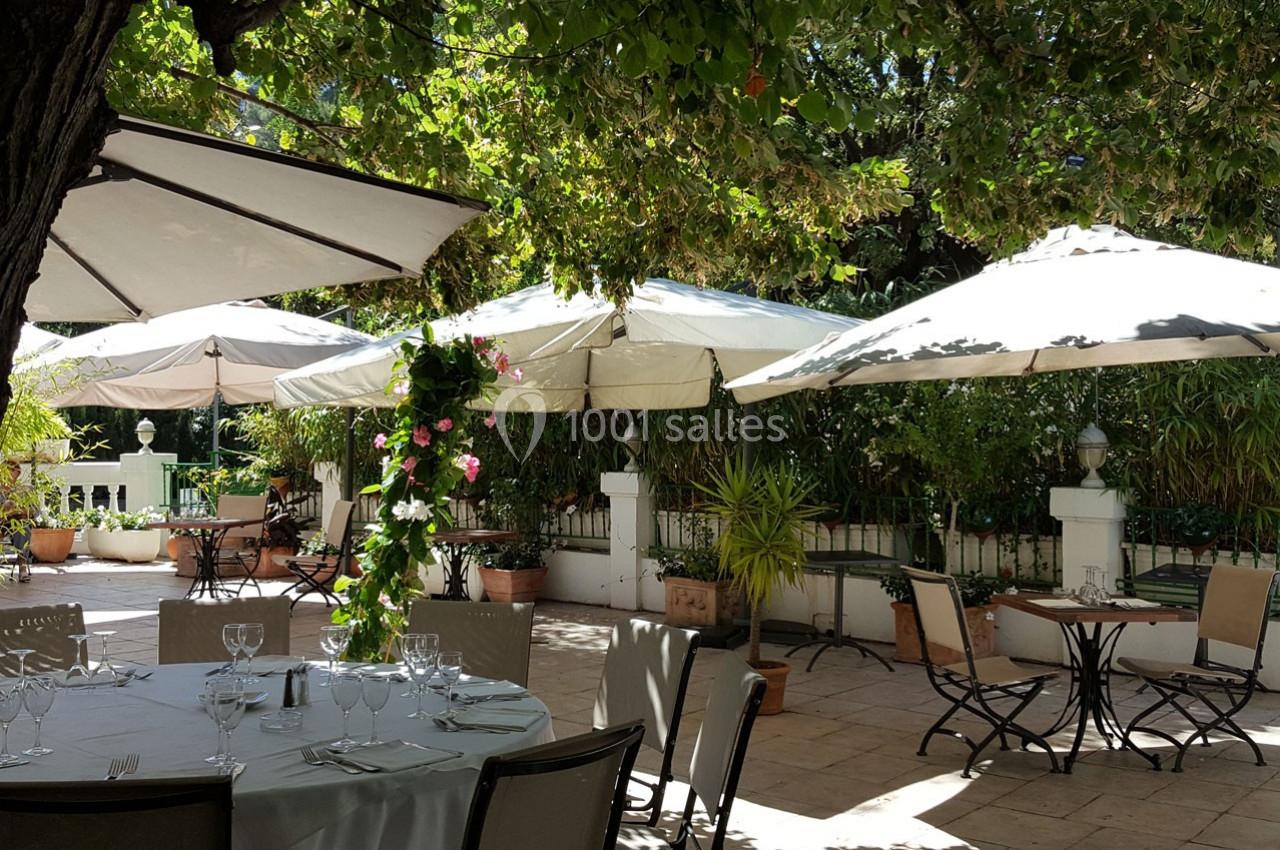 Terrasse ombragée avec tables dressées, chaises et parasols, entourée de verdure et de plantes en pots.