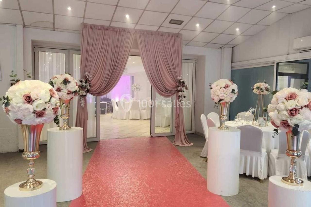 Salle de réception décorée avec des fleurs blanches et roses, tapis rouge menant à des tables et chaises blanches.