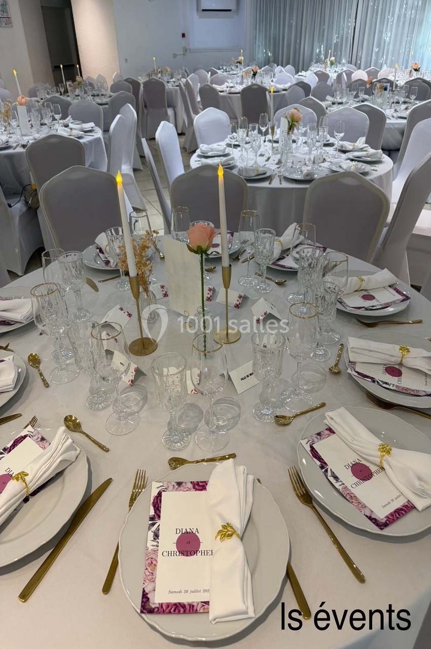 Salle de réception avec tables rondes dressées, décorées de bougies, fleurs et menus personnalisés.