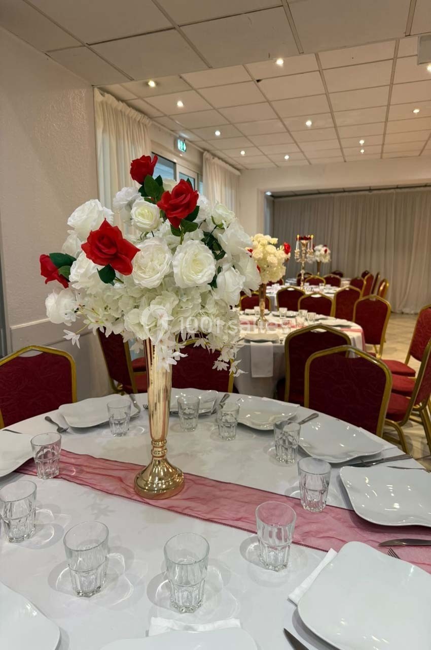 Salle de réception décorée avec des tables dressées, ornées de centres de table composés de roses rouges et blanches.