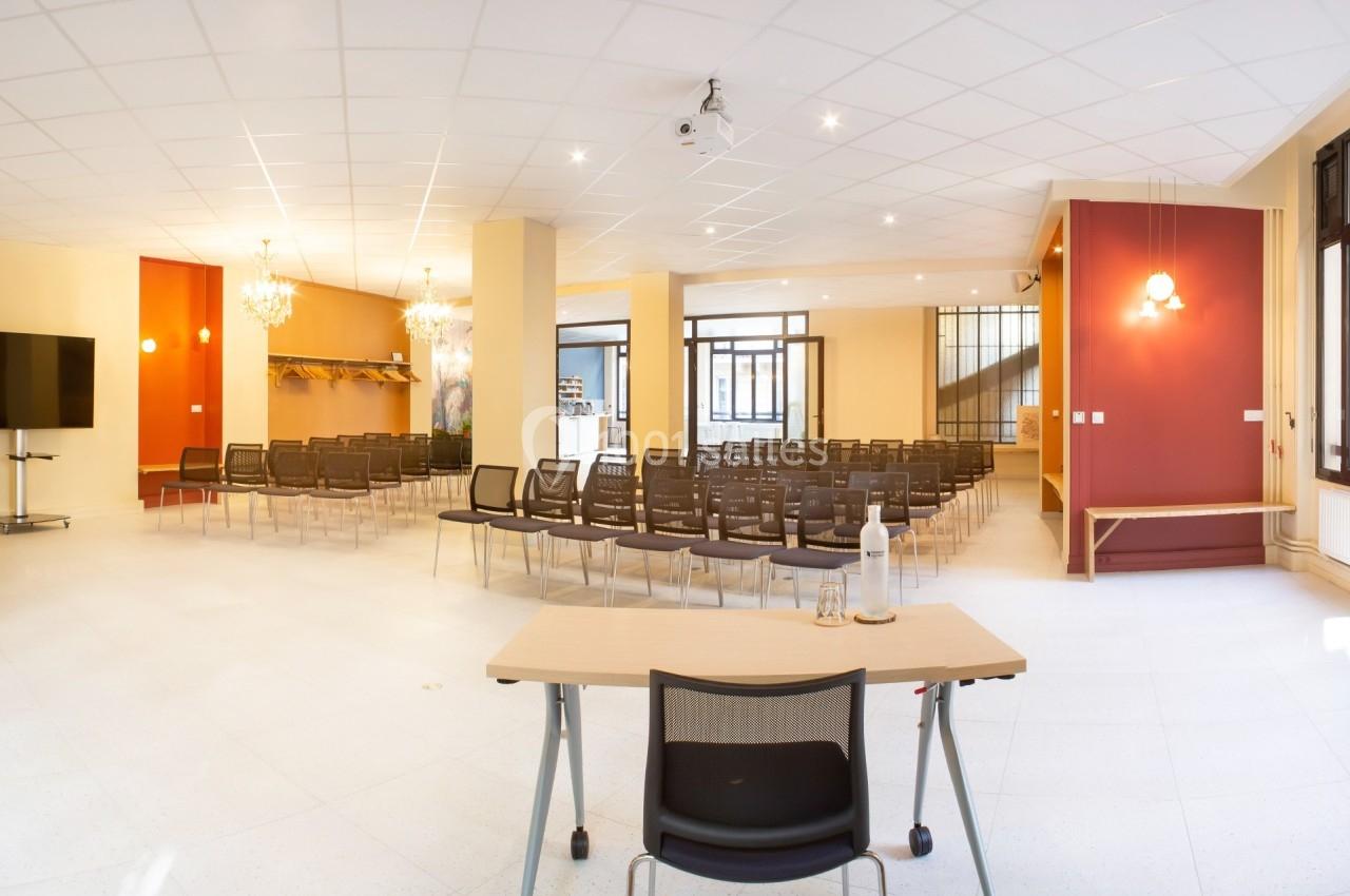 Salle lumineuse avec rangées de chaises, un bureau au premier plan et des lustres suspendus au plafond.