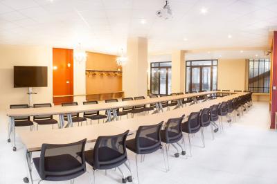 Salle de réunion lumineuse avec tables alignées, chaises noires, bouteilles d'eau et étagères en arrière-plan.