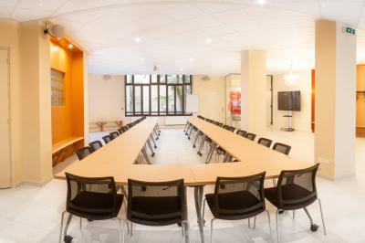 Salle de réunion lumineuse avec tables alignées, chaises noires, bouteilles d'eau et étagères en arrière-plan.