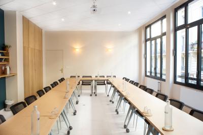 Salle de réunion lumineuse avec tables alignées, chaises noires, bouteilles d'eau et étagères en arrière-plan.