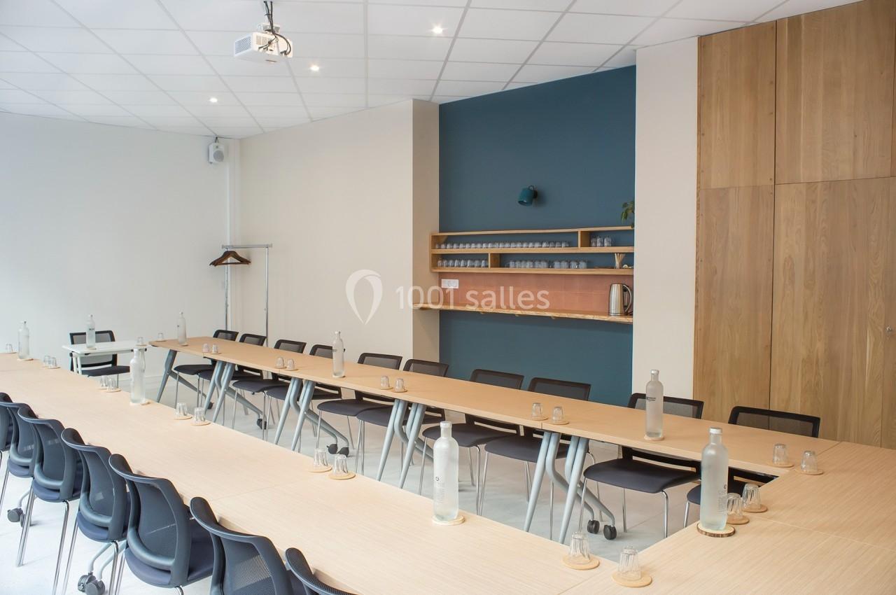 Salle de réunion lumineuse avec tables en bois disposées en U, chaises noires et étagères murales en arrière-plan.