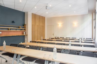Salle de réunion lumineuse avec tables alignées, chaises noires, bouteilles d'eau et étagères en arrière-plan.