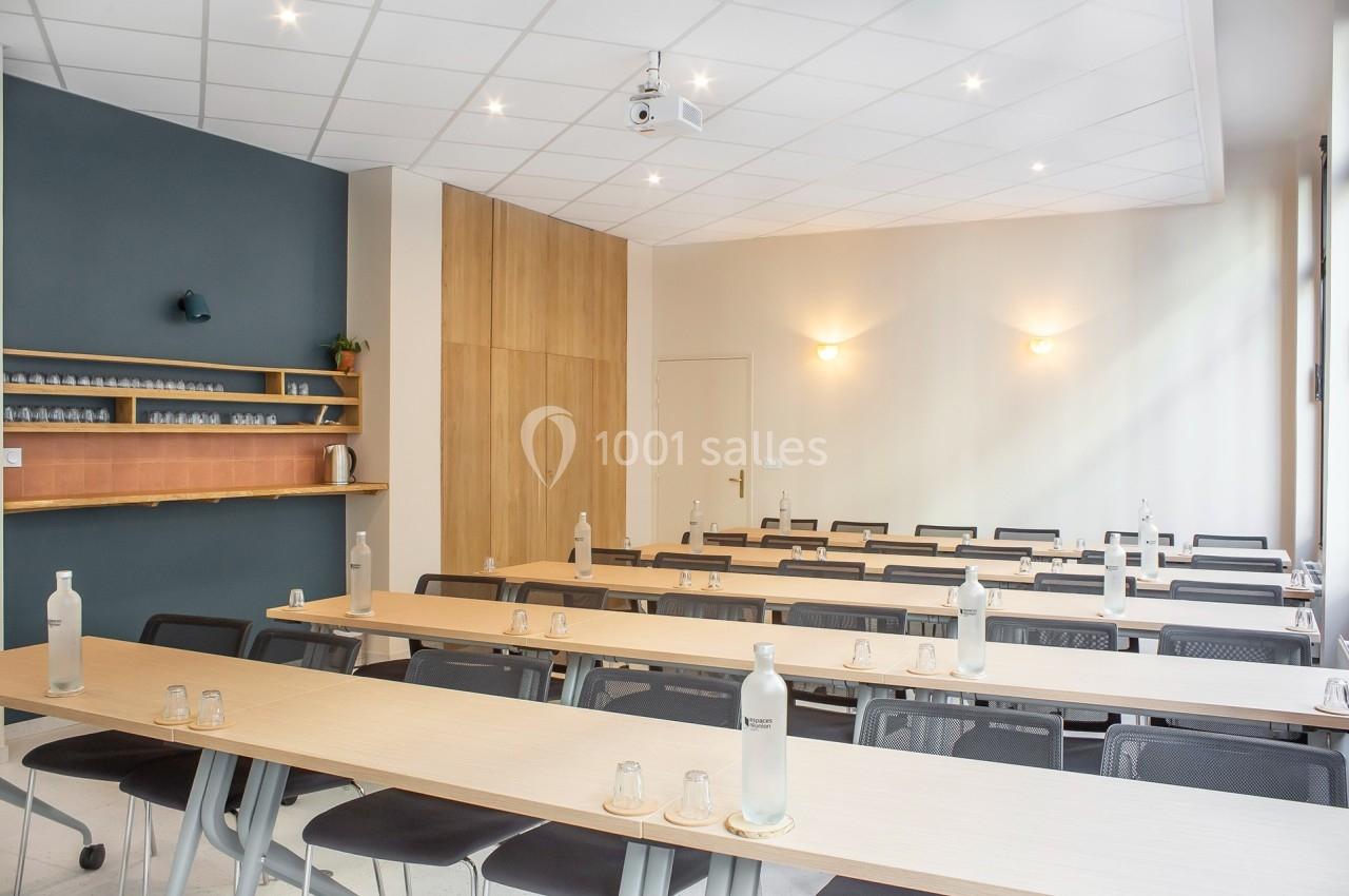 Salle de réunion lumineuse avec des tables alignées, des chaises noires et des bouteilles d'eau disposées.