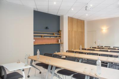 Salle de réunion lumineuse avec tables alignées, chaises noires, bouteilles d'eau et étagères en arrière-plan.