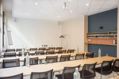 Salle de réunion lumineuse avec tables alignées, chaises noires, bouteilles d'eau et étagères en arrière-plan.