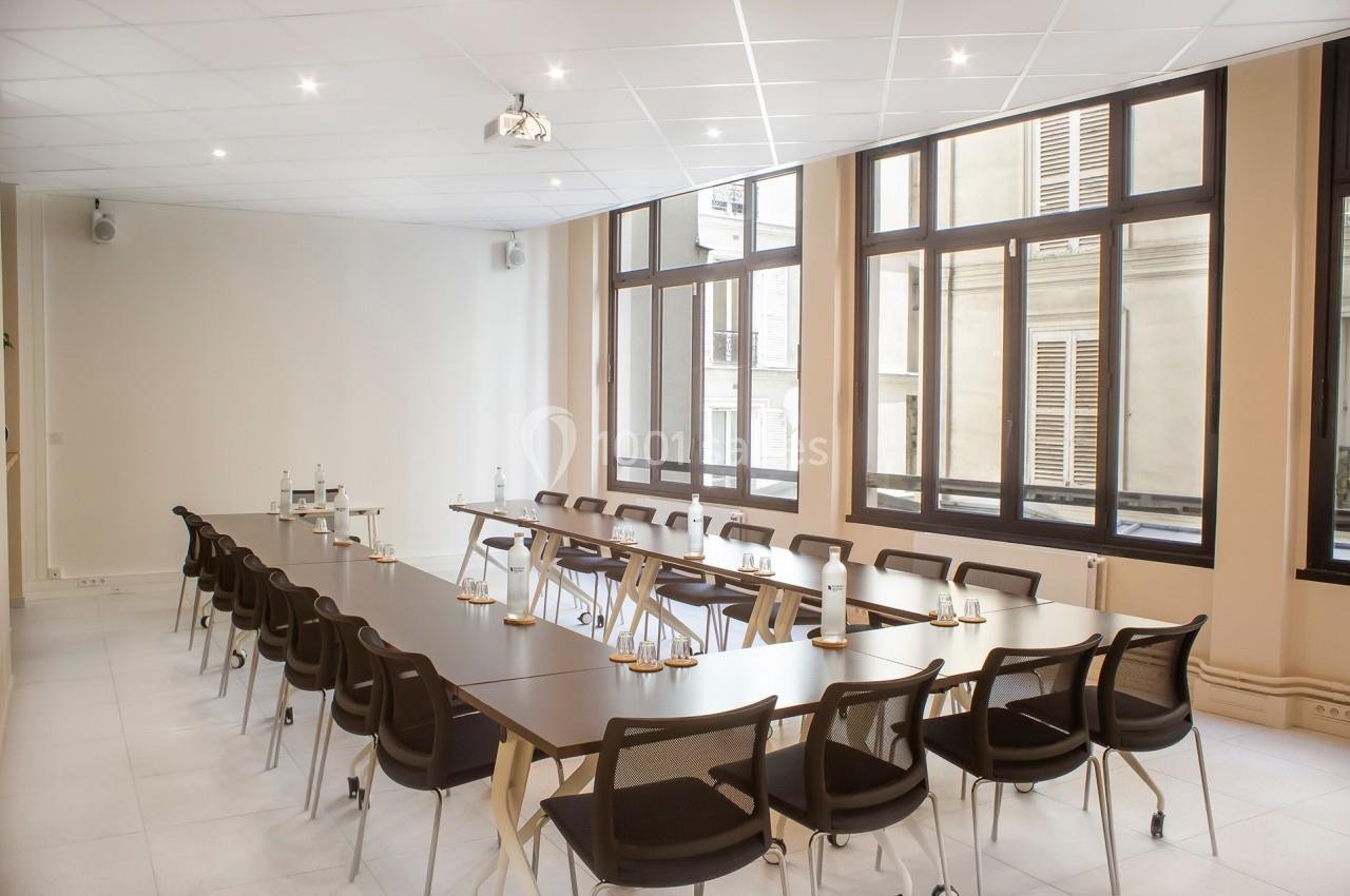 Salle de réunion lumineuse avec de grandes fenêtres, des tables disposées en U et des chaises noires.