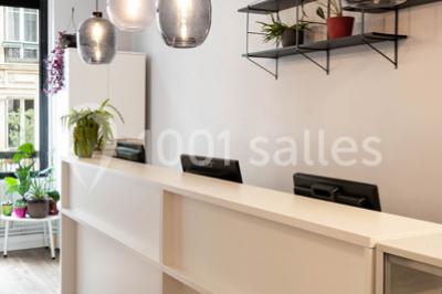 Salle de réunion lumineuse avec tables alignées, chaises noires, bouteilles d'eau et étagères en arrière-plan.