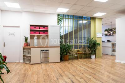 Salle de réunion lumineuse avec tables alignées, chaises noires, bouteilles d'eau et étagères en arrière-plan.