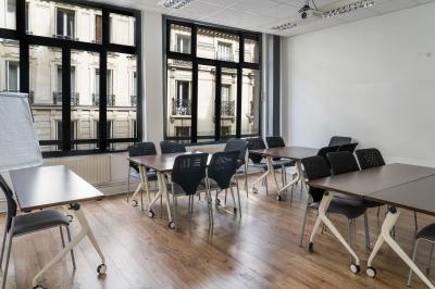 Salle de réunion lumineuse avec tables alignées, chaises noires, bouteilles d'eau et étagères en arrière-plan.