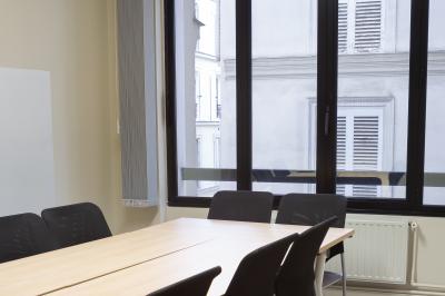 Salle de réunion lumineuse avec tables alignées, chaises noires, bouteilles d'eau et étagères en arrière-plan.
