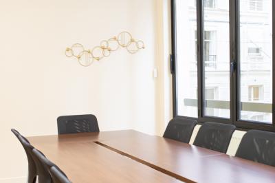 Salle de réunion lumineuse avec tables alignées, chaises noires, bouteilles d'eau et étagères en arrière-plan.