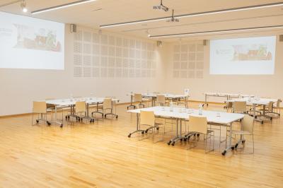 Salle de réunion lumineuse avec tables alignées, chaises noires, bouteilles d'eau et étagères en arrière-plan.