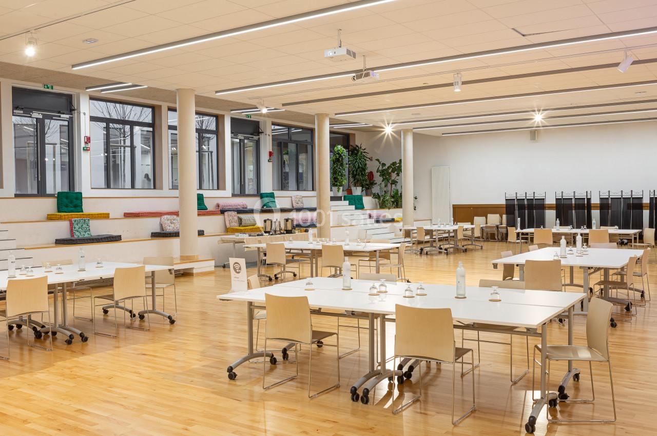 Salle lumineuse avec tables et chaises en bois, espace de travail ou réunion, gradins avec coussins colorés en arrière-plan.