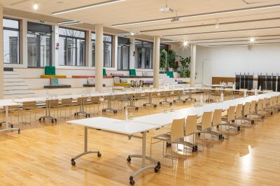 Salle de réunion lumineuse avec tables alignées, chaises noires, bouteilles d'eau et étagères en arrière-plan.