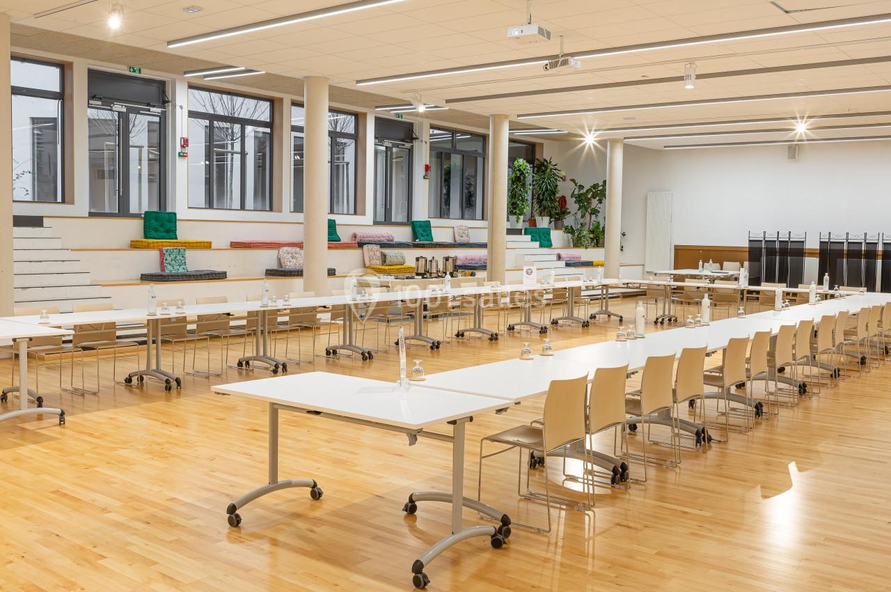 Salle lumineuse aménagée pour une réunion, avec tables blanches, chaises alignées et espace de détente en arrière-plan.