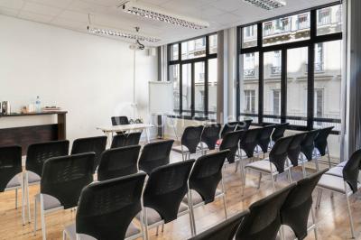 Salle de réunion lumineuse avec tables alignées, chaises noires, bouteilles d'eau et étagères en arrière-plan.
