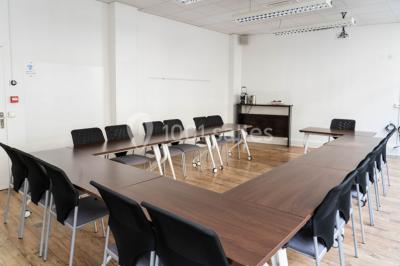 Salle de réunion lumineuse avec tables alignées, chaises noires, bouteilles d'eau et étagères en arrière-plan.