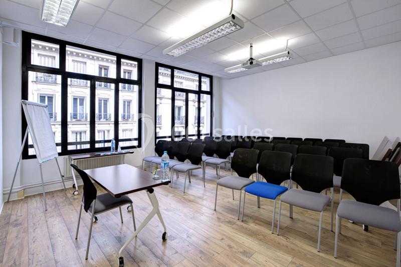 Salle de réunion lumineuse avec chaises alignées, table, paperboard et grandes fenêtres donnant sur des immeubles.