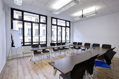 Salle de réunion lumineuse avec tables alignées, chaises noires, bouteilles d'eau et étagères en arrière-plan.