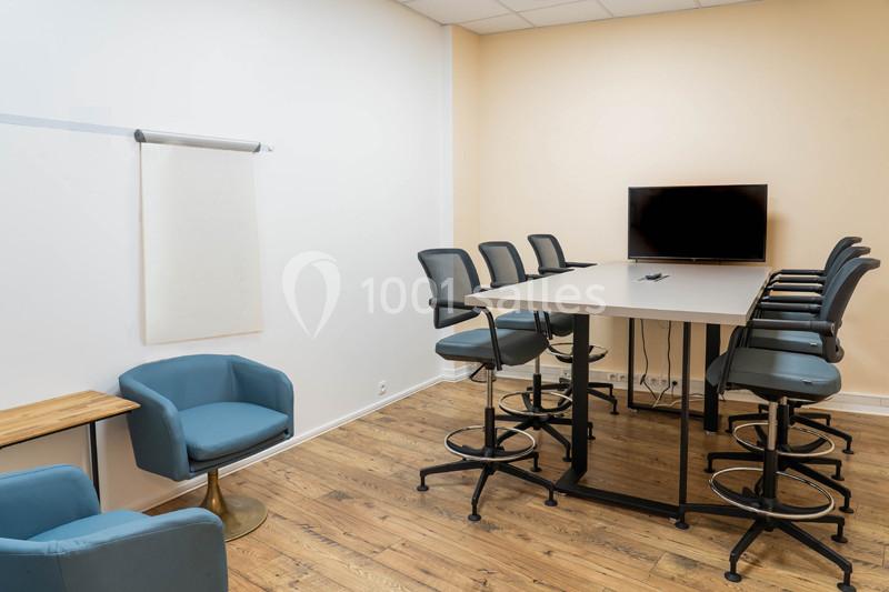 Salle de réunion avec table haute, chaises hautes, écran mural, tableau blanc et fauteuils bleus sur sol en bois.