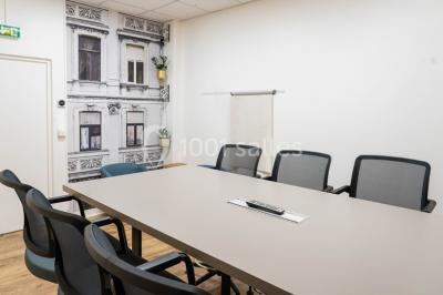 Salle de réunion lumineuse avec tables alignées, chaises noires, bouteilles d'eau et étagères en arrière-plan.