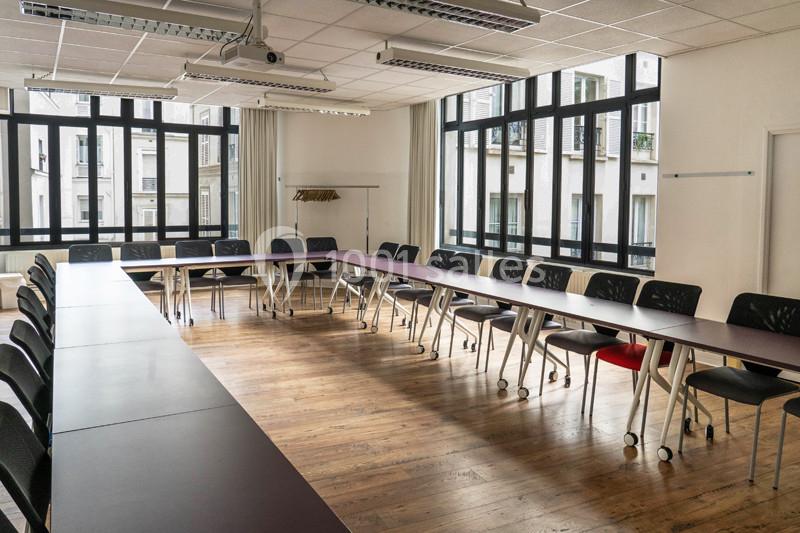 Salle de réunion lumineuse avec de longues tables en U, des chaises noires et un sol en parquet.