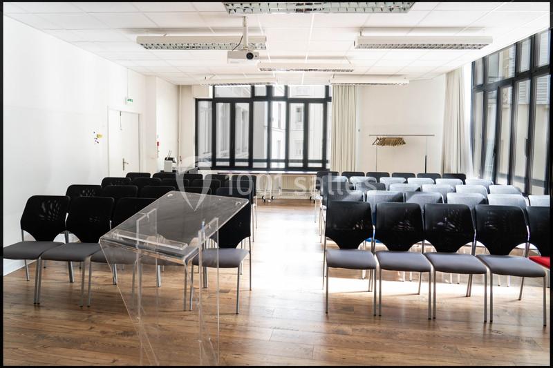 Salle de conférence lumineuse avec rangées de chaises noires, pupitre transparent et grand écran au plafond.