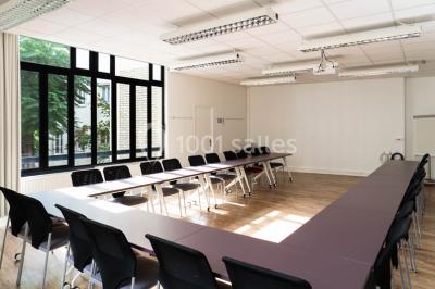 Salle de réunion lumineuse avec tables alignées, chaises noires, bouteilles d'eau et étagères en arrière-plan.