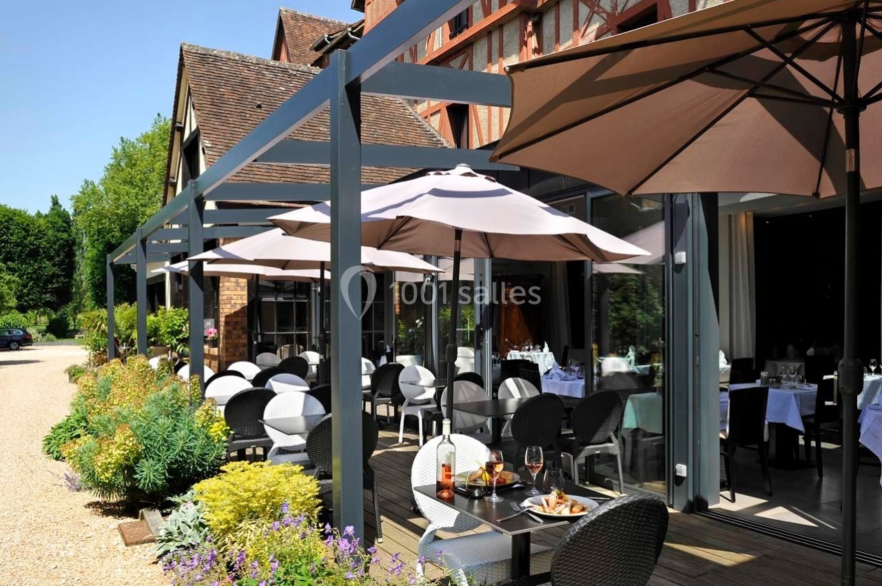 Terrasse d'un restaurant avec tables dressées, chaises en osier et parasols, entourée de végétation et d'un bâtiment en…