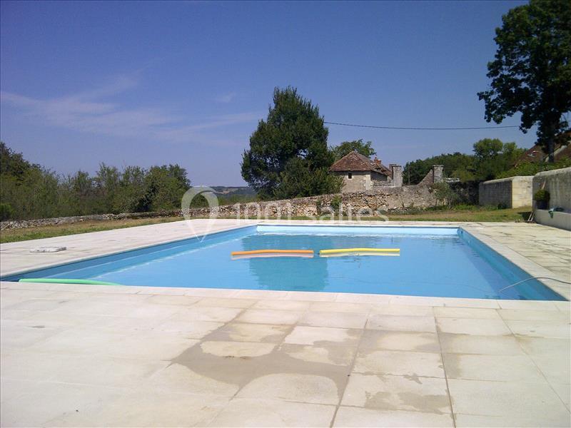 Piscine extérieure rectangulaire entourée d'une terrasse en pierre, avec des arbres et des bâtiments en arrière-plan.