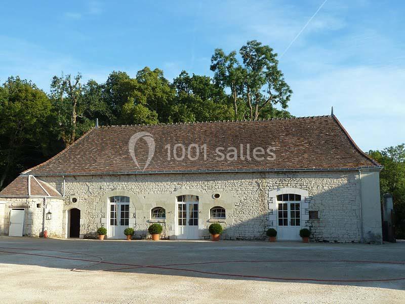 Bâtiment en pierre avec toit en tuiles, grandes fenêtres blanches et pots de fleurs, entouré de végétation.
