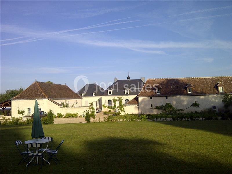 Cour d'une propriété avec bâtiments en pierre, pelouse, table et chaises sous un parasol, ciel dégagé.