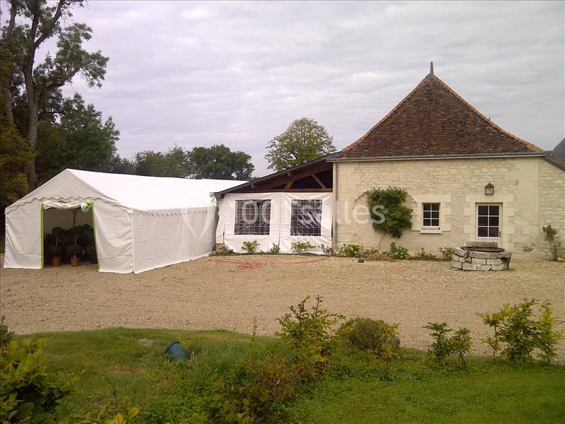Bâtiment en pierre avec toit en pente, tente blanche attenante et puits en pierre dans une cour gravillonnée.