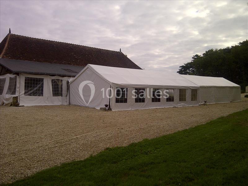 Grande tente blanche installée sur un sol en gravier, à côté d'un bâtiment en pierre avec un toit en tuiles.