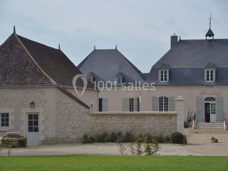 Bâtiments en pierre avec toits en ardoise, fenêtres à volets et cour gravillonnée, entourés de verdure.