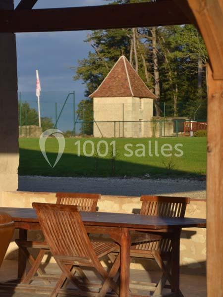 Table en bois avec chaises sur une terrasse, vue sur un jardin verdoyant et un petit bâtiment en pierre au fond.