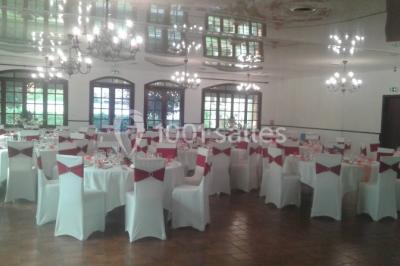 Salle de réception lumineuse avec tables dressées, chaises blanches, chandeliers et décorations florales.