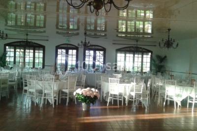 Salle de réception lumineuse avec tables dressées, chaises blanches, chandeliers et décorations florales.