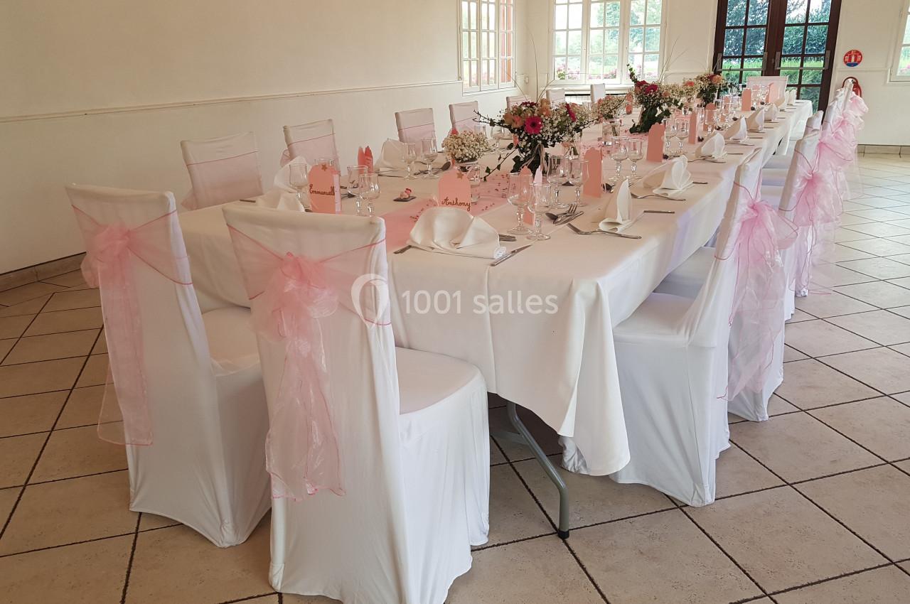 Table dressée pour un événement, avec nappes blanches, chaises décorées de nœuds roses et centre de table fleuri.