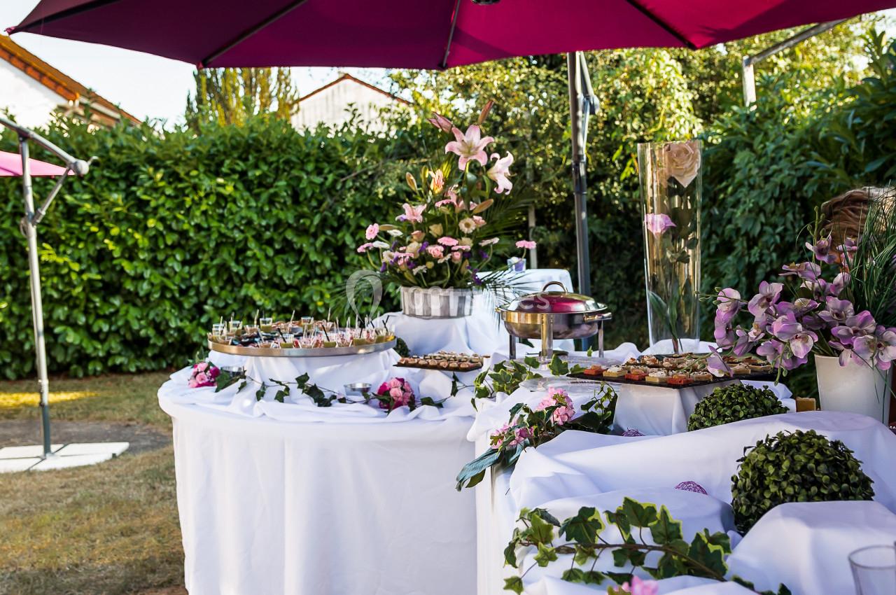 Buffet en extérieur avec nappes blanches, fleurs roses et parasols violets, entouré de verdure.