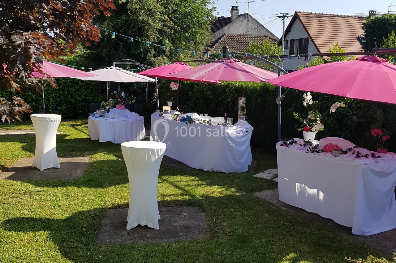 Jardin aménagé pour une réception avec tables décorées, nappes blanches et parasols roses, entouré de verdure.