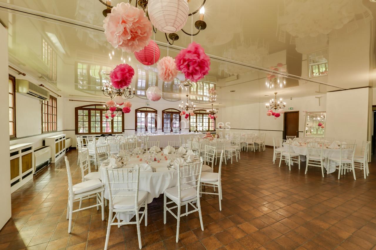 Salle de réception décorée de lanternes et pompons roses, tables rondes dressées avec nappes blanches et chaises assorties.