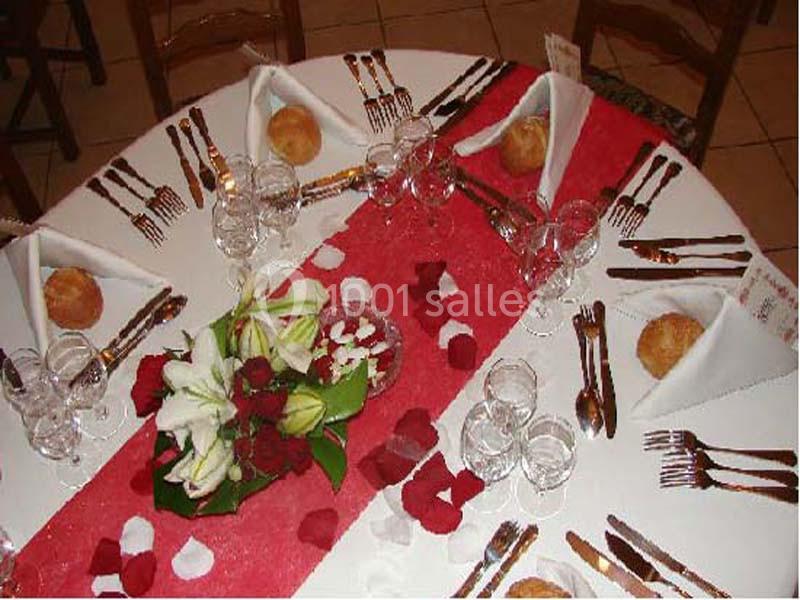 Table ronde dressée avec nappes blanches, chemin de table rouge, fleurs, pétales, couverts et petits pains.