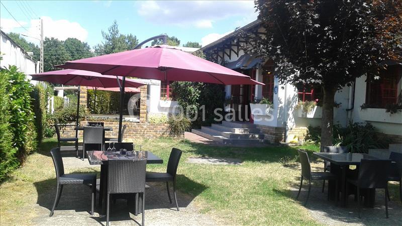 Terrasse extérieure avec tables, chaises en rotin et parasols rouges, entourée de verdure et d'un bâtiment en arrière-plan.